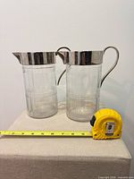 Front view of both glass pitchers showing metal lids, handles, and height tape for scale