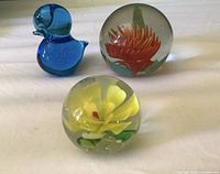 Group shot showing blue bird figurine, orange flower paperweight, yellow flower paperweight