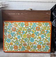 Two folded tables leaning against wall: floral child table in front, faux wood table behind