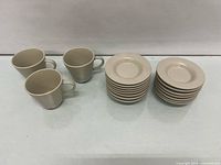 Stack of 15 beige ceramic saucers beside three matching mugs