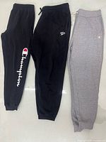 Three pairs of men’s sweatpants laid flat: black Champion, black Slazenger, grey Champion