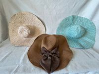 Front view of three woven sun hats—beige, aqua, and brown with bow—displayed on white cloth
