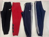 First four sweatpants laid out: black Puma, red, navy with script logo, navy with white stripes