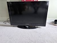 Front view of Dynex 32 inch TV on base with remote positioned at stand, power cord trailing behind