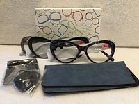 three pairs +2.50 reading glasses in front of patterned box with cases and pouches