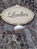 Front view of cream metal wall sign with "Ladies" text and ceramic knob