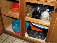 Kitchen lower cabinet open with plastic mixing bowls, trays, canisters, and a manual citrus juicer stored on shelves