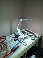 Desk setup showing white Ott light with rectangular head illuminated, visible Halloween eyeball lights, tangled cords including red tubing, remote control on table.