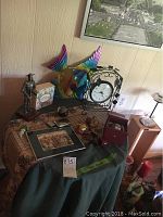 Full view of wood octagon table with various decorative items including metal peacock, knight figurine, clock, art print, small items grouped on a cloth covering the table.