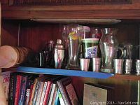 Wide view of shelf with assorted metal shot glasses, drink shakers, glass pitchers, and branded plastic cups with Rolling Rock and Margaritaville logos.