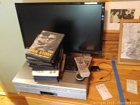 Front view showing silver Apex VHS/DVD player, stack of DVDs, two remote controls on top, and RCA flat screen TV behind.