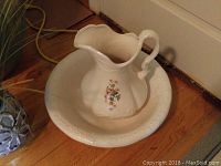 White ironstone water pitcher with floral decoration inside matching ironstone basin with embossed patterns, placed on wood floor.