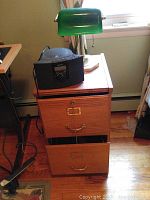 Wood file cabinet with three drawers, brass handles and label holders, one drawer open, with green glass desk lamp and Sony Dream Machine radio on top