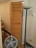 Wooden room divider shutter standing behind a white floor fan with three blades; fan shows some wear and small crack on top cage; hardwood floor visible