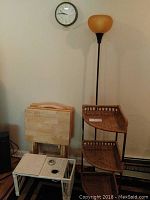 Standing wicker shelf with two baskets, tall floor lamp with orange shade, round wall clock on white wall, and small folding wooden snack tables.