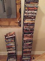 Two tall uneven stacks of DVDs on the floor against wall, showing a mix of titles and warped cases.
