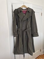 Full view of trench coat hanging, showing olive exterior, belt, double-breasted design, epaulettes, cuff straps