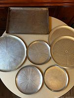 Six round pizza trays (various sizes, two perforated) and one rectangular baking sheet displayed on tabletop
