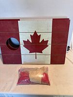 Front view of one cornhole board showing Canada flag design with hole and plastic bag containing red and tan beanbags; second board edge visible behind