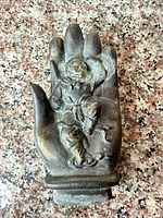 Front view of cast metal hand supporting seated figure, showing overall design, color and patina