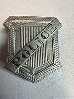 Front view of silver-tone police badge pin showing raised "POLICE" banner and textured shield design