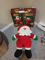 Two boxed musical Santas and plush Santa doll displayed on floor