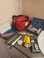 Overview of lot on countertop showing red first aid bag, blue Auto Kit, flashlights, LED light, Braun thermometer package, three-hole punch and strap