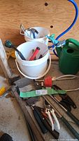 Assorted garden tools including buckets with hammers, pruners, loppers, bow saw frame and pickaxe visible