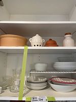 Overview of cupboard with top and middle shelves showing terracotta casserole, garlic keeper, butter dish, ceramic bottle, glassware and stacked dinnerware