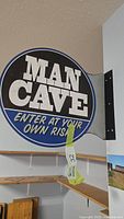 Front view of round steel sign displaying 'MAN CAVE ENTER AT YOUR OWN RISK' text, mounted on wall bracket.