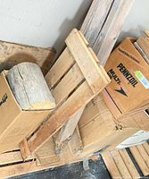 Pile of wood offcuts: split log round, pallet slats, cardboard boxes, long boards leaning against wall