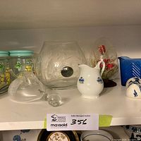 Shelf showing glass jars, etched bowl, spout barometer, creamer, egg paperweight, blue dish, souvenir dish