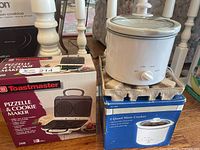 Both appliances with their retail boxes; pizzelle maker boxed, slow cooker displayed on packing insert atop its box