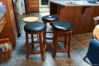 Three bar stools with wooden legs and black vinyl padded round seats, and one smaller wooden stool with no cushion in a kitchen setting on tile floor.