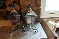 Two metal and glass lanterns side by side on a stone surface with red candles inside, with curled metal stands.