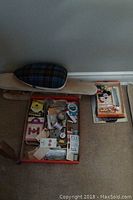 View of the box filled with assorted sewing supplies and a wooden ironing or tailor's board with a plaid cover beside it.