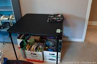 Black folding card table shown fully assembled with boxes stored underneath.