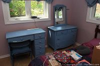Blue dresser with attached mirror and blue desk arranged under windows, folding chair in front of desk, showing overall set condition including paint wear and scratches