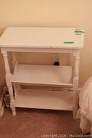 Front view of the white painted wooden side table with built-in angled magazine rack on middle shelf showing table surface, legs and bottom shelf.