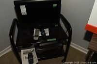 Photo of black Sanyo 18-inch TV on black metal stand with JVC VCR and Philips DVD player below, with manuals and remotes on top shelf.