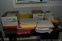 Stacks of legal size photo copy paper, colored paper, continuous computer paper boxes, and envelopes arranged on a table.