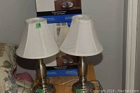 Two brushed steel finish touch table lamps with white fabric bell-shaped shades, displayed on a wooden surface with tapered bases and touch sensors.