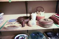 Photo showing the full collection displayed on a shelf, featuring multiple handmade pottery items including plates, vases, and a lidded casserole dish.