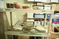 Photo of assorted handmade pottery bowls, plates, and trays on a plastic shelving unit, showing variety of small to medium items in different earth tones.