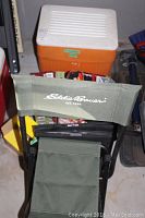 Eddie Bauer folding stool with green fabric seat and backrest, orange and white Coleman cooler in background along with some bags.