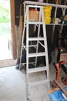 Photo of a silver aluminum 6-foot step ladder standing in a garage with various tools and storage items in the background, showing the ladder's front.