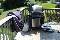 Full view of Broil King gas BBQ grill on wooden deck with a propane tank beside it and BBQ accessory tools laid next to the grill. Grill has folding side shelves and a cover draped on railing.