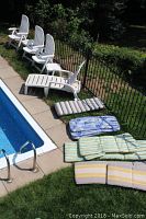 Six white plastic lawn chairs, two small tables, one lounge chair and assorted cushions arranged near a poolside and lawn area.