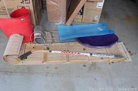 Wooden toboggans with plastic snow slides laid out on the floor, alongside a hockey stick and red bin.