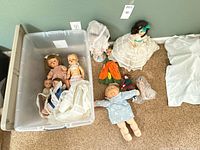 Overview of lot showing bin with five dolls, additional two large dolls, extra clothing